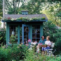 a man and woman sitting on a bench in front of a garden shed with flowers