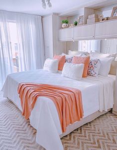 a bed with white sheets and orange pillows in a bedroom next to a large window