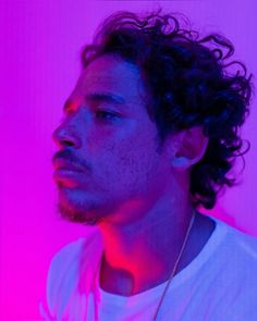 a man with curly hair and a white t - shirt looks off into the distance