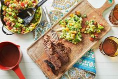 a wooden cutting board topped with meat and vegetables next to two bowls of salads