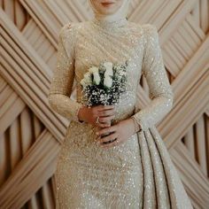 a woman in a white dress holding flowers and standing next to a wall with wood paneling