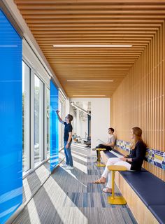 three people sitting on benches in an office with blue walls and windows, one person pointing at something