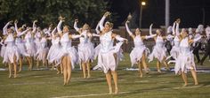 a group of young women dressed in white dance on the field at night with their hands up