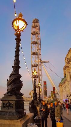 people are standing on the sidewalk next to a street light and ferris wheel at dusk