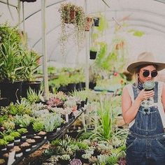 a woman wearing overalls and a hat taking a selfie in a greenhouse