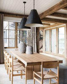 a dining room table with chairs and two vases on top of it in front of large windows