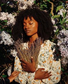 a woman standing in front of flowers with her eyes closed and hands folded over her chest