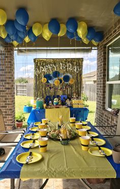 a table set up for a party with blue and yellow balloons hanging from the ceiling