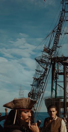 two men standing next to each other in front of an old fashioned pirate ship on the ocean