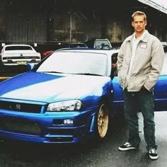 a man standing next to a blue car in a parking lot with other cars behind him