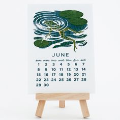 a calendar with a frog on it sitting on a wooden easel in front of a white background