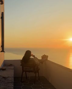 a woman sitting on a chair looking out at the ocean while the sun is setting