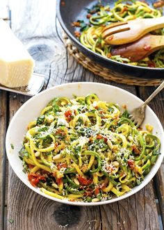 two bowls filled with pasta and vegetables on top of a wooden table next to cheese