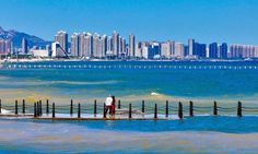 two people standing on a pier in front of the ocean with city skyline behind them