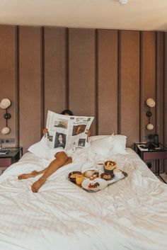 a person laying on a bed reading a newspaper and having breakfast with coffee, croissants and fruit