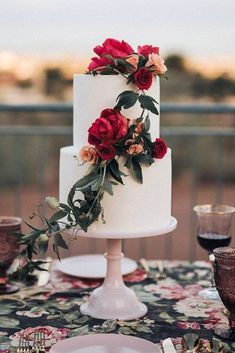 a white wedding cake with red flowers and greenery sits on a floral tablecloth