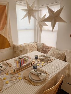 the table is set with plates, cups and utensils in front of paper stars hanging from the window