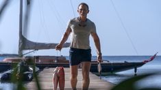 a man walking on a dock with a fish in his hand and a sailboat in the background