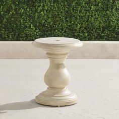 a white vase sitting on top of a cement floor next to a hedge covered wall