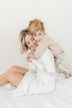 a woman holding a small child on top of a white bed in front of a white wall