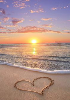 a heart drawn in the sand on a beach at sunset, with the sun setting behind it