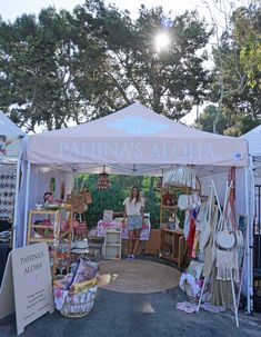 a woman standing in front of a tent with items for sale on the ground and trees behind her