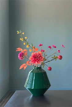 a green vase filled with lots of flowers on top of a table next to a wall