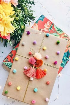 two wrapped gifts with pom - poms on them and flowers in the background