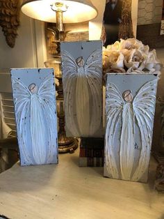 three white angel paintings sitting on top of a table next to a lamp and flowers