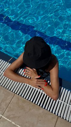 a woman sitting on the edge of a swimming pool wearing a black hat and red nail polish