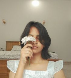 a woman holding a flower in front of her face and looking at the camera while sitting on a bed