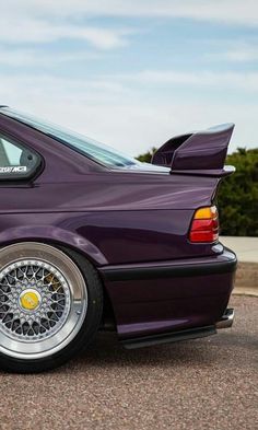 the back end of a purple car parked in a parking lot next to trees and bushes