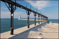 a long pier with a light house in the distance and snow on the ground next to it