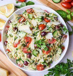 a white bowl filled with pasta salad next to sliced cucumbers and lemon wedges