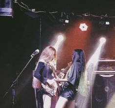 two women standing on stage playing guitars and singing into microphones with lights behind them