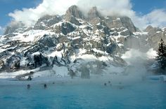 people are swimming in a blue pool with snow on the mountains