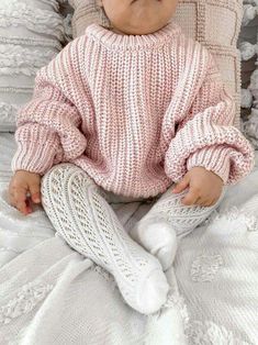 a baby wearing a pink sweater and white leggings sitting on top of a bed