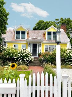 a white picket fence in front of a yellow house with sunflowers and bushes