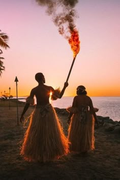 two women dressed in grass skirts holding torches on the beach at sunset with palm trees and water in the background