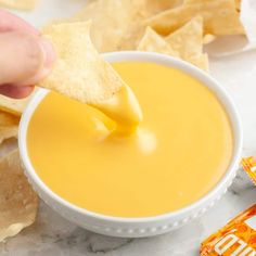 a person dipping tortilla chips into a white bowl filled with orange liquid and topped with cheese