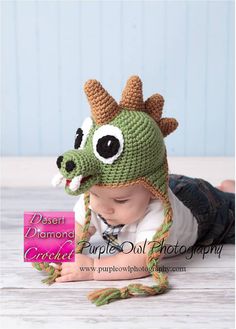 a baby wearing a crocheted dragon hat laying on the floor with a pink sign