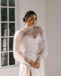 a woman standing in front of a window wearing a white dress with sheer sleeves and high slit