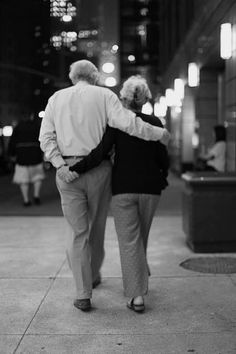 an older couple walking down the sidewalk in front of a building at night, back to back