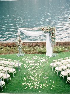 an outdoor ceremony setup with white chairs and flower petals on the grass near the water