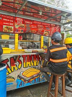 some people are sitting at a food stand and one is wearing a yellow safety helmet