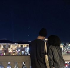 a man and woman standing on top of a roof looking at the city lights in the distance