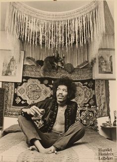 a black and white photo of a man sitting on a bed in front of a tapestry