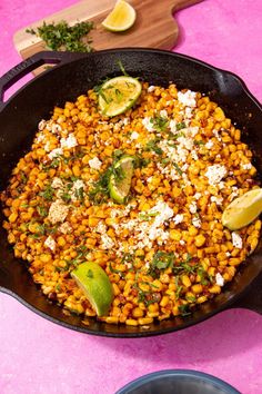 a skillet filled with corn and topped with feta cheese, lime wedges and cilantro