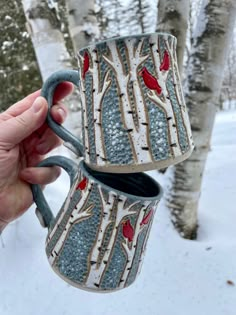 someone holding two mugs in the snow with trees on them and one has red birds painted on it