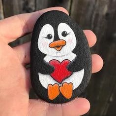 a hand holding a painted rock with a penguin holding a heart in it's paws